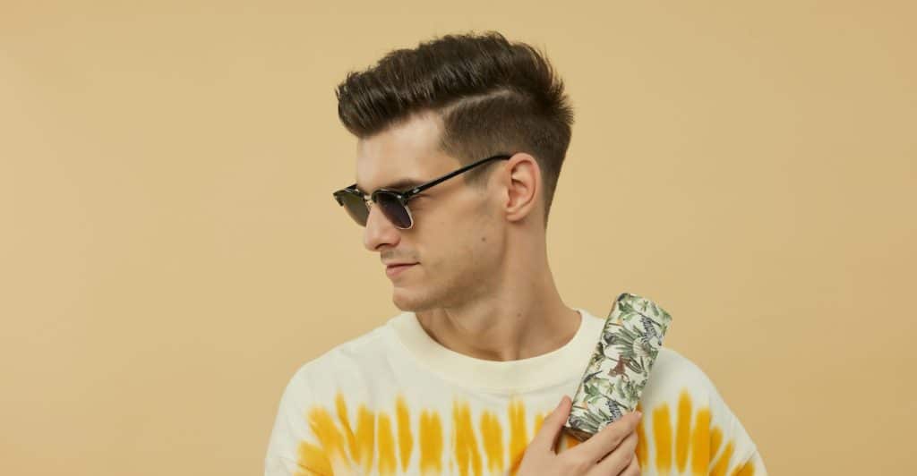 A man with styled hair and sunglasses, wearing a tie-dye shirt, holding sunglasses on his shoulder.