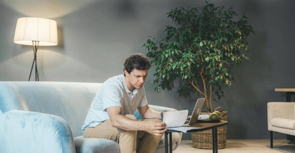 A man with brown hair is sitting on a gray couch in a room with dark walls, looking down at a laptop on his lap and holding a piece of paper in his left hand, with several white pendant lights hanging from the ceiling.