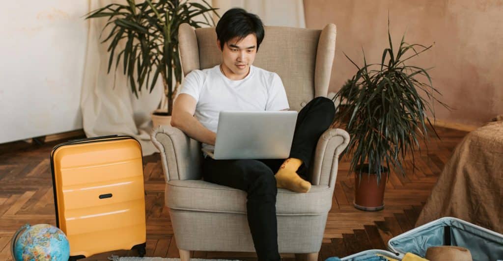 A man sitting in an armchair with a laptop on his lap, surrounded by luggage and a globe, looking at the screen.