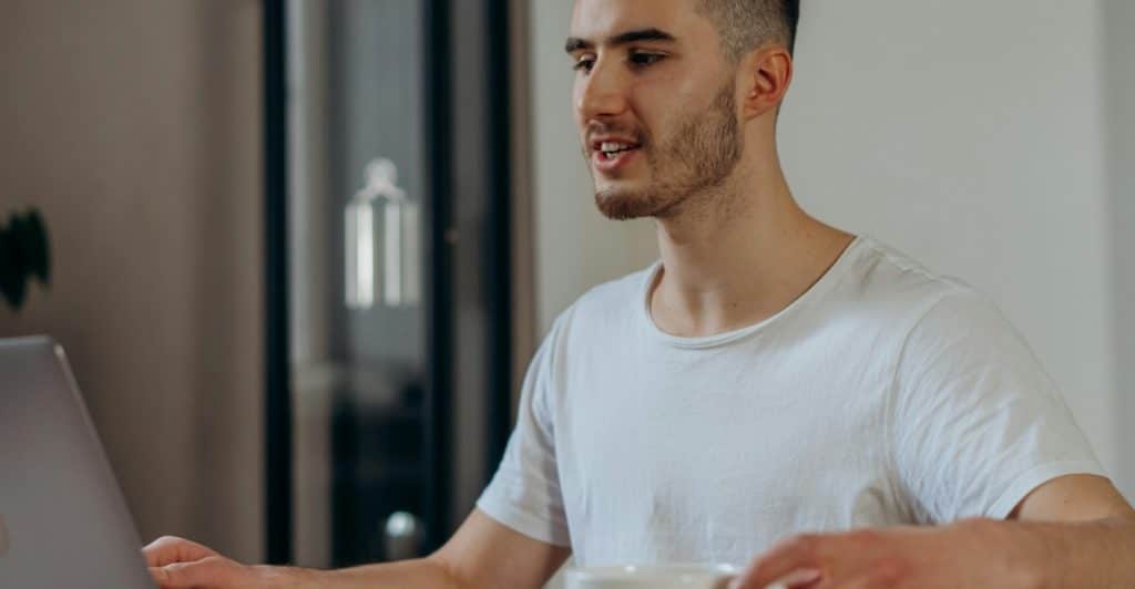 A man smiling and looking at a laptop while holding a coffee cup and saucer.