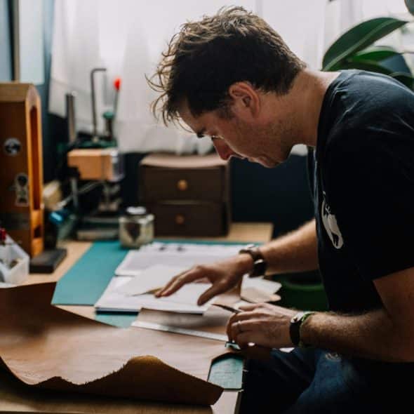 A man with wavy brown hair wearing a black t-shirt focused on working with a piece of leather on a desk filled with tools and materials.