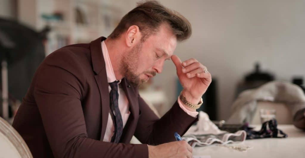 A man in a suit jacket and tie, with a pen in hand, looking down thoughtfully while writing at a table.