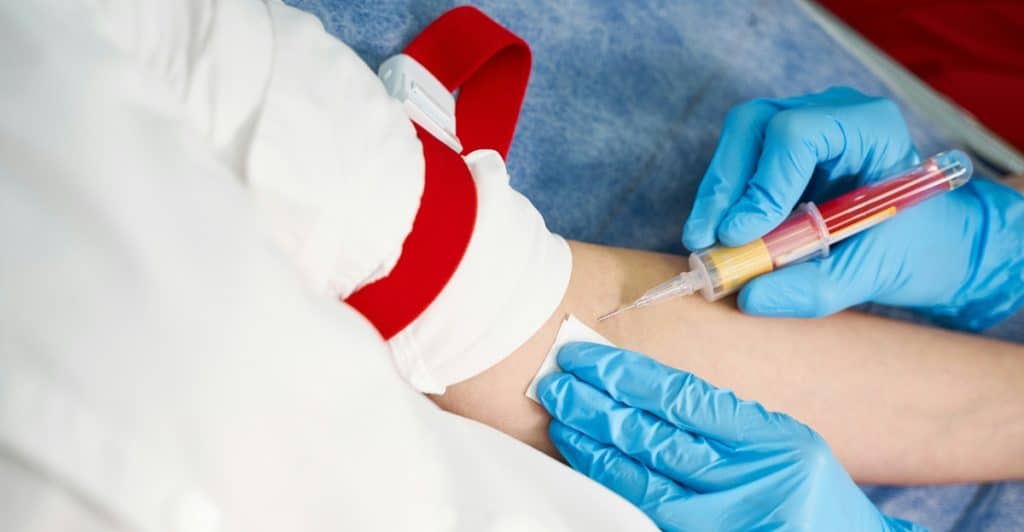 A medical professional in blue gloves is drawing blood from a person's arm using a needle and a blood collection tube.