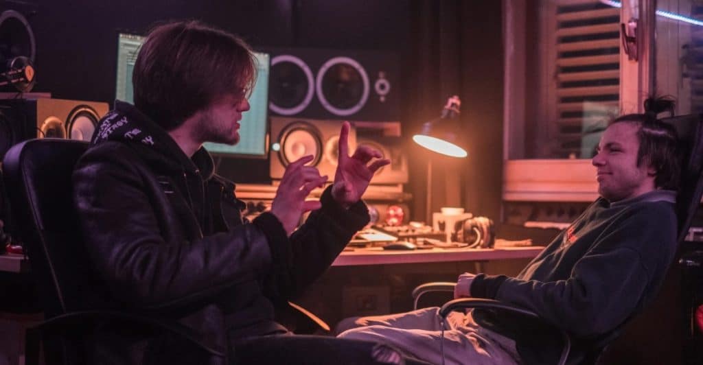 Two men sitting in chairs in a recording studio, one gesturing while speaking to the other.