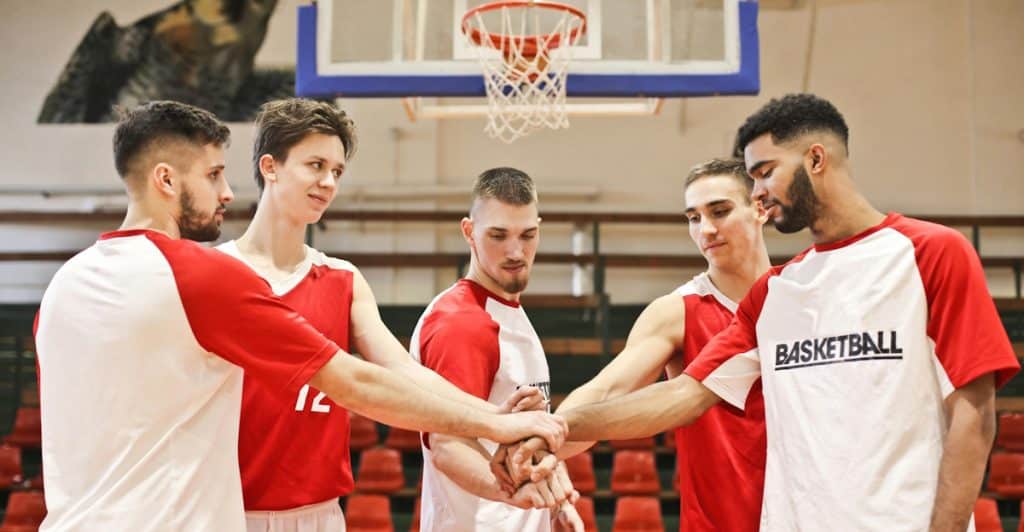 Five men in red and white basketball uniforms are standing on a court with their hands stacked in the center.