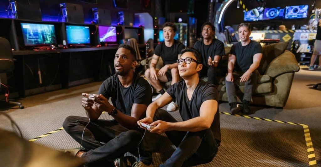 Four men in a dimly lit room with multiple screens, with two men in the foreground sitting on the floor playing video games.