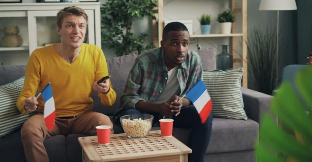 Two men sitting on a couch holding French flags and watching television with popcorn and red cups on a coffee table.