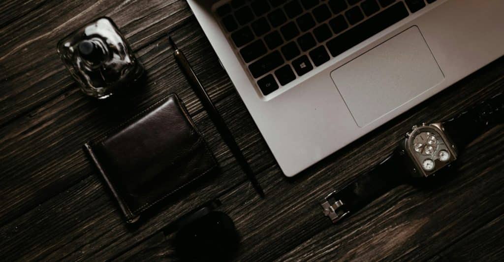 A watch, a wallet, and a bottle of cologne are arranged on a dark wooden surface next to a laptop.