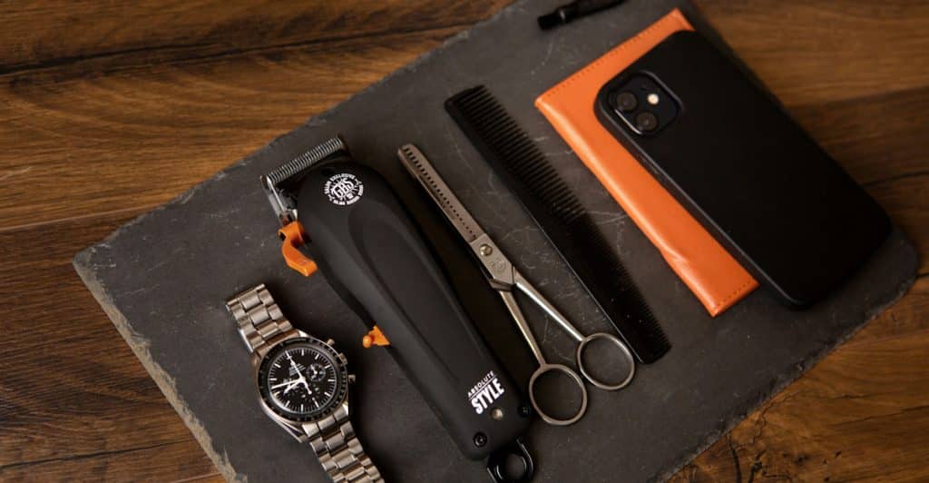 A collection of men's grooming accessories including a watch an electric trimmer scissors and a comb laid out on a dark slate surface.