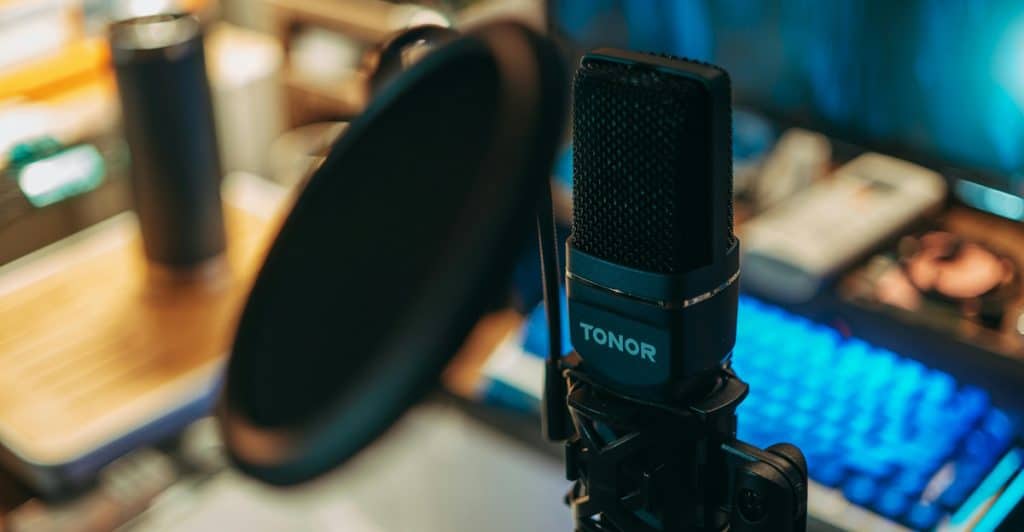 A close-up of a black condenser microphone on a stand, with a gaming keyboard featuring blue backlighting visible in the background.