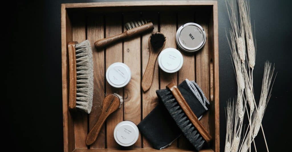 An overhead view of various wooden grooming brushes and tins of wax arranged in a wooden tray next to stalks of wheat.