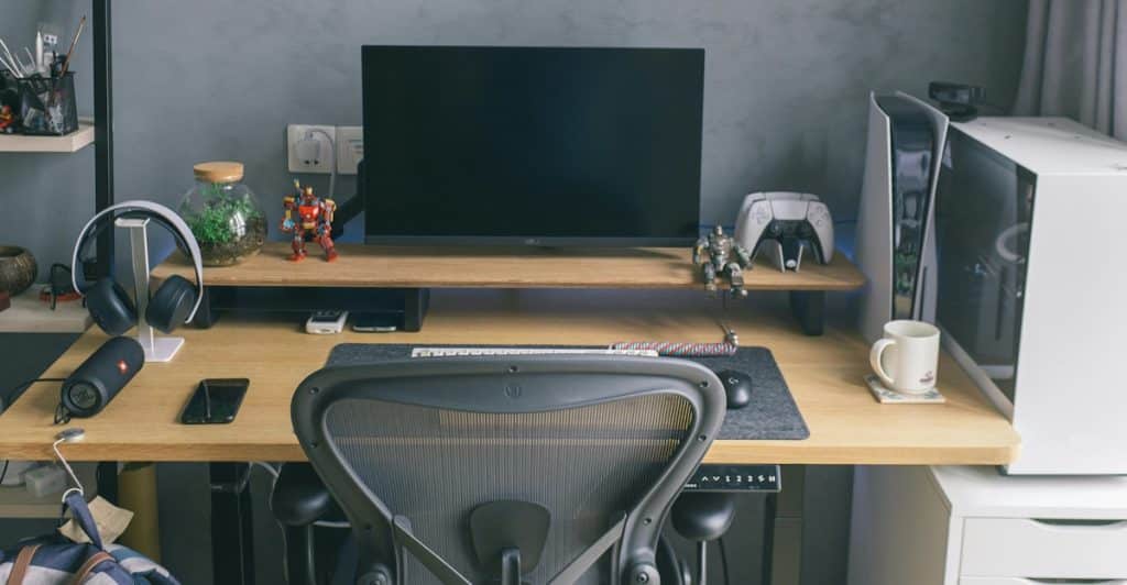 A well-organized gaming desk features a large monitor on a wooden riser, a white desktop PC, headphones, and various accessories, all against a gray wall.