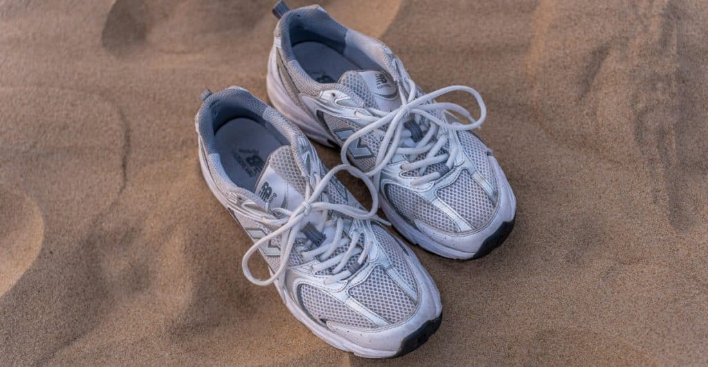 A pair of grey New Balance 990 series sneakers with white laces resting on sand.