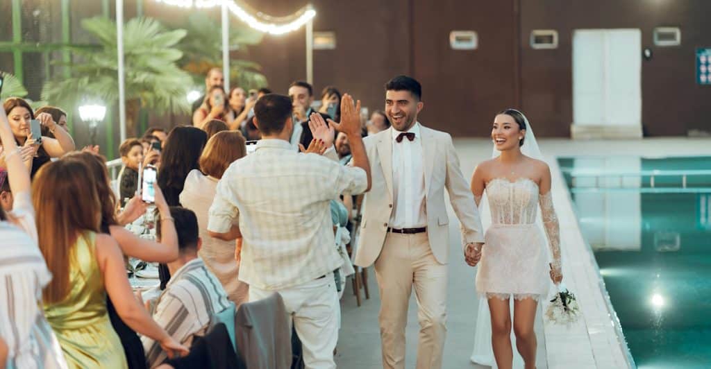 A smiling newlywed couple, the man in a light suit and the woman in a short white dress, are walking hand-in-hand past a crowd of people at an outdoor evening event with a swimming pool to their right.