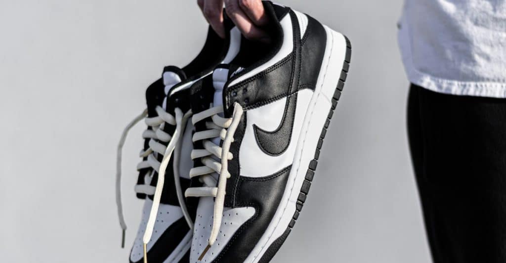 A person holding a pair of black and white Nike Dunk Low sneakers by the laces.