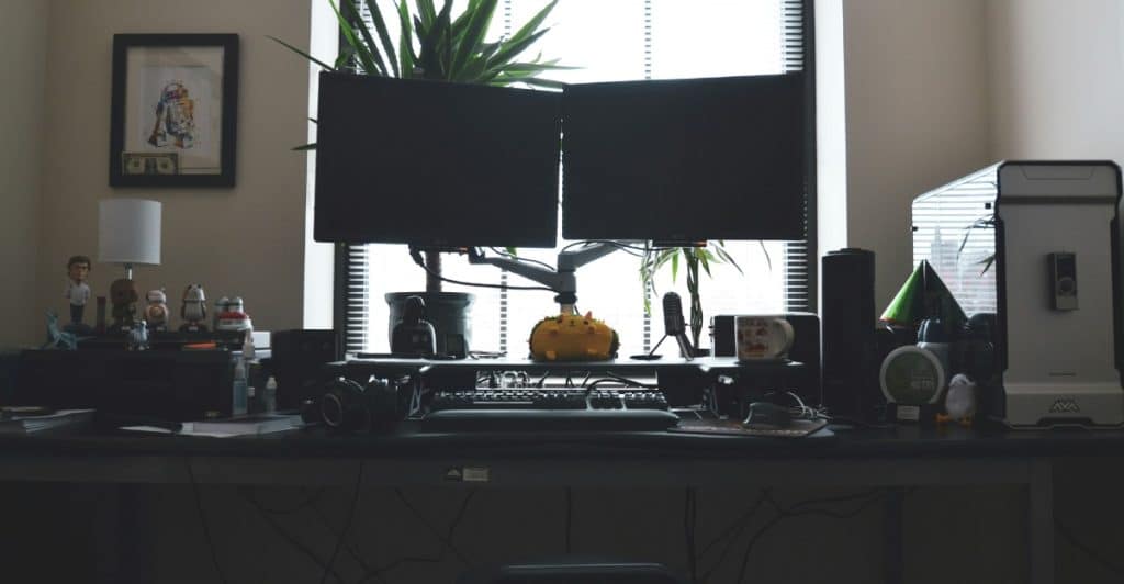 An office desk with two monitors, various tech gadgets, and plants in front of a window with blinds