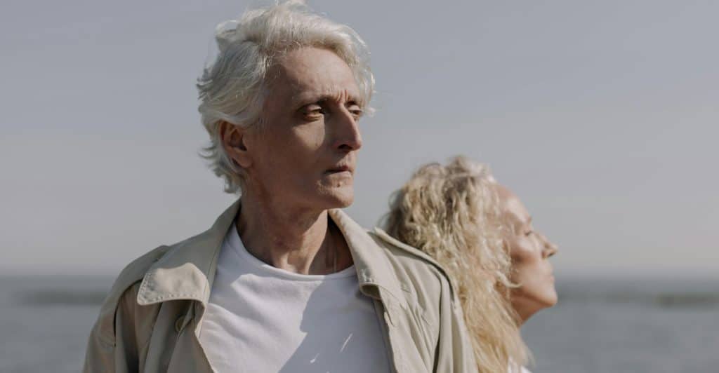 An older man with white hair and a beige coat looking seriously forward, standing back-to-back with an older woman with blonde curly hair and a white shirt, both near a body of water.