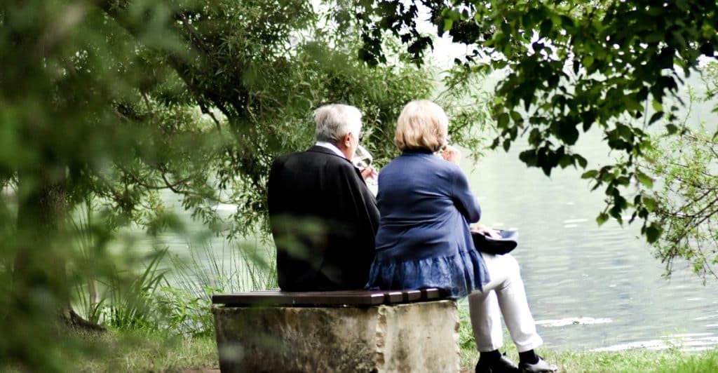 An older couple sitting on a bench next to a body of water, seen from behind.