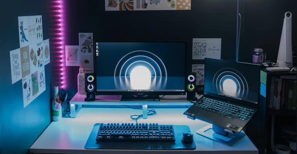 An organized desk setup with a monitor, laptop, speakers, and a vertical LED light strip along the wall, demonstrating clean cable management.