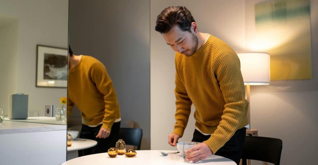 A man in a yellow sweater standing in a minimalist room, organizing small items on a white round table next to a large mirror.
