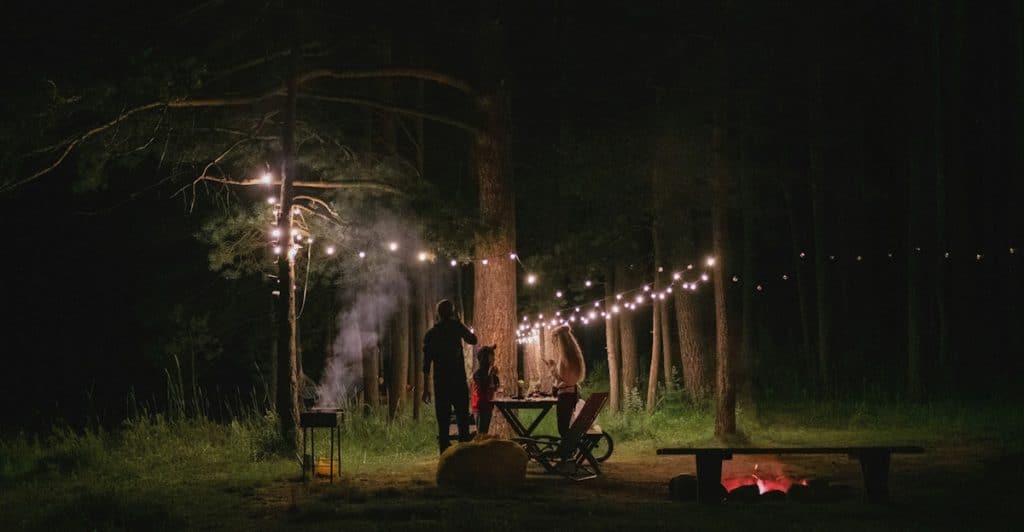 Two people silhouetted against a dark forest background, setting up string lights and tending to a grill at an outdoor night gathering.