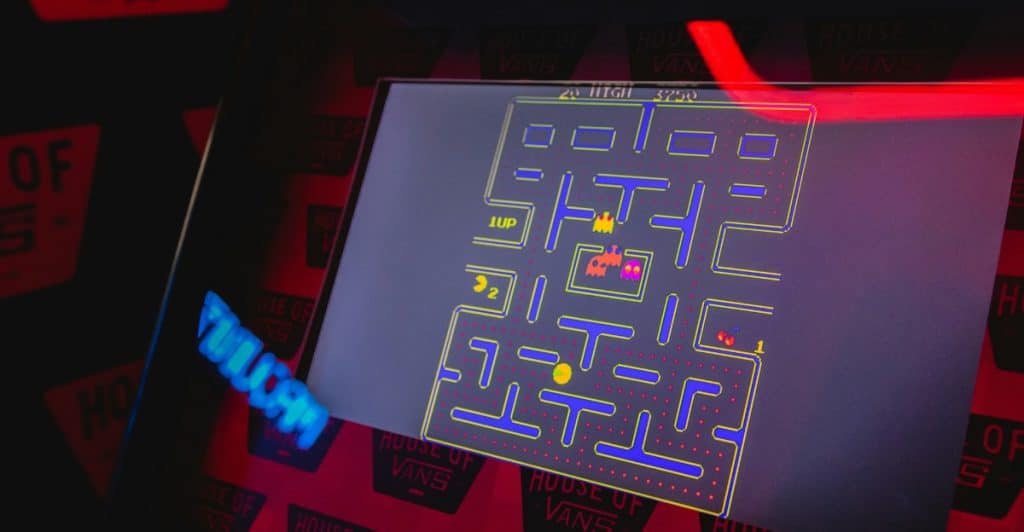 A close-up of a Pac-Man arcade game screen showing the maze, Pac-Man, and ghosts with a "House of Vans" neon sign in the background.