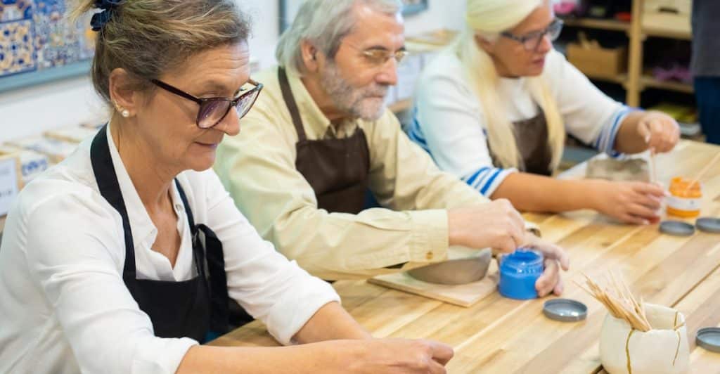 Three elderly people, two women and one man, wearing aprons and eyeglasses, actively engaged in painting their molded pots at a wooden table in an art studio.