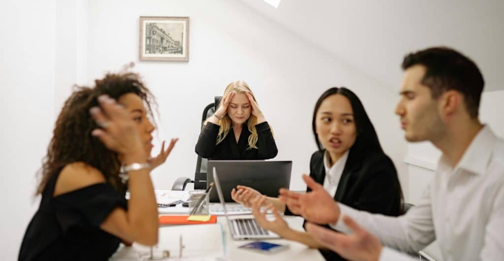 Two women and a man sitting at a table in an office, appearing to be in a heated discussion with a woman holding her head in the background.
