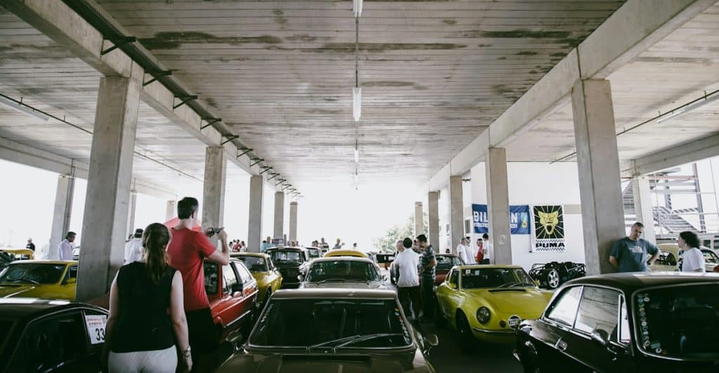 People gathered around classic cars in a large, open garage or parking structure.