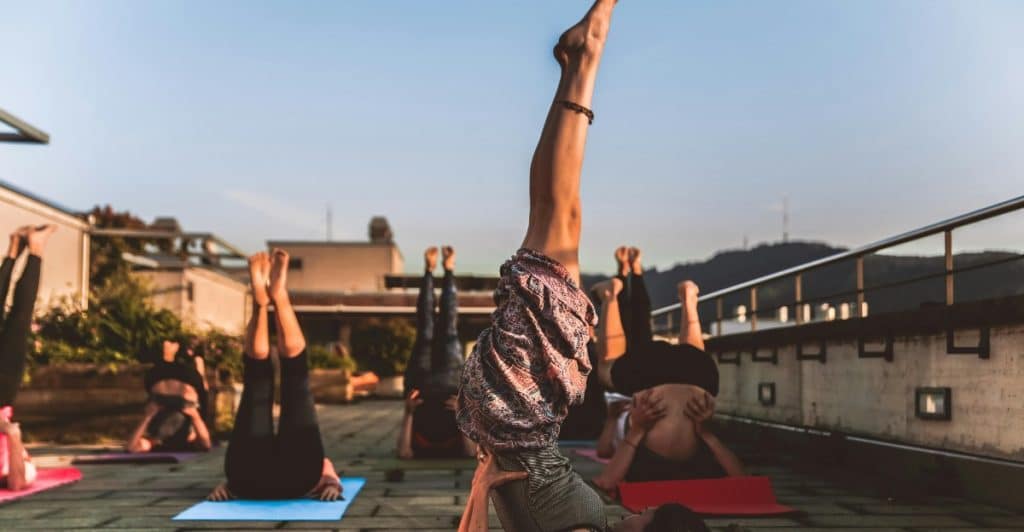 Multiple people performing a yoga pose where they are balancing on their shoulders with their legs extended upwards, on yoga mats outdoors.
