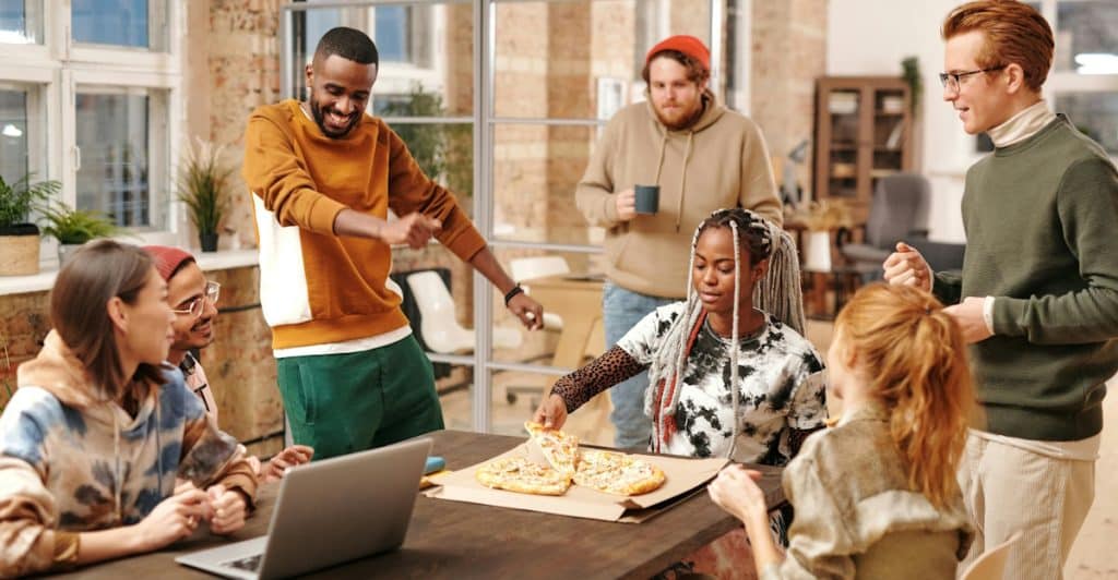 A group of diverse individuals gathered around a table with pizza, some standing and some sitting, engaged in conversation.