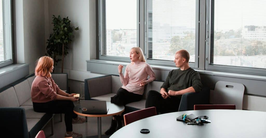 Three people, two women and one man, sitting around a table in a modern office or counseling setting, engaged in conversation near large windows.