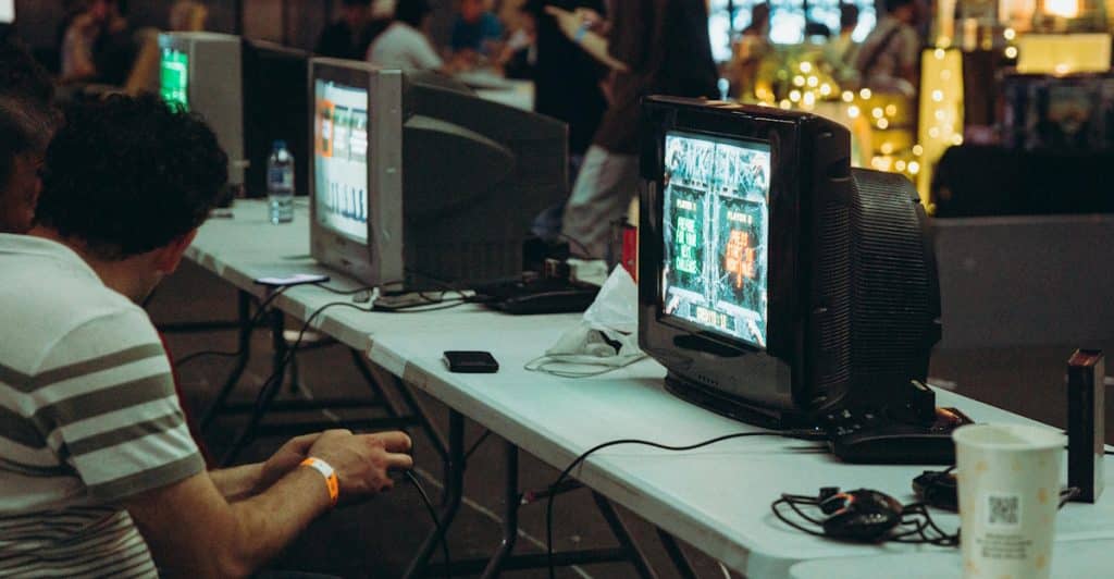 Several people are seated at tables, playing classic video games on large, box-style CRT televisions at what appears to be a gaming event.