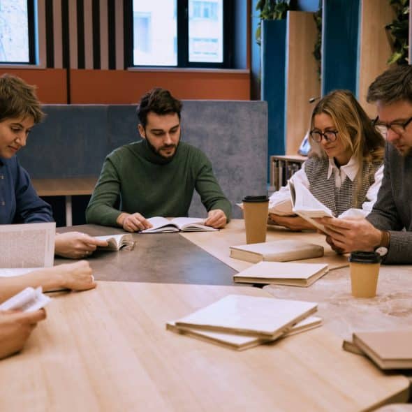 A group of people sitting around a wooden table, some holding open books and others with coffee cups nearby.