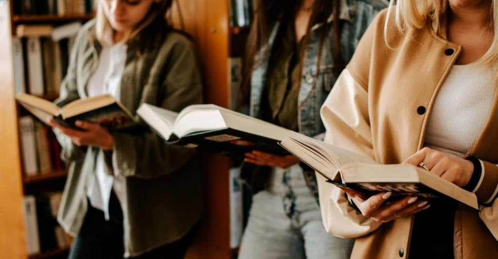 Three people standing in a library, each holding an open book and reading