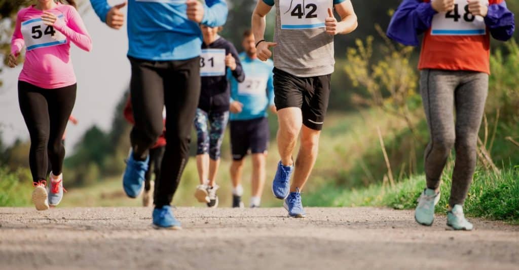 Several people running outdoors on a paved path, participating in a race with numbers pinned to their shirts