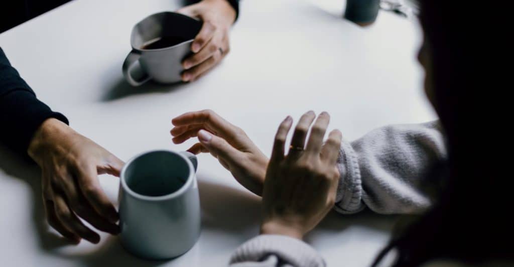 Two people with mugs on a white table, one with hands clasped and the other's hands visible, engaged in conversation.