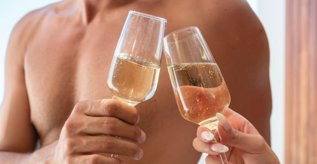 Two people toast with champagne flutes, one bare-chested and the other with manicured nails.