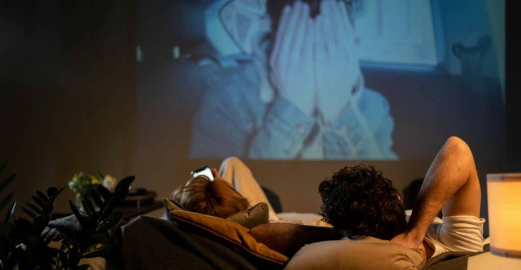 Two people lying on a couch, one looking at a phone, while an emotional scene from a movie is projected onto a wall.