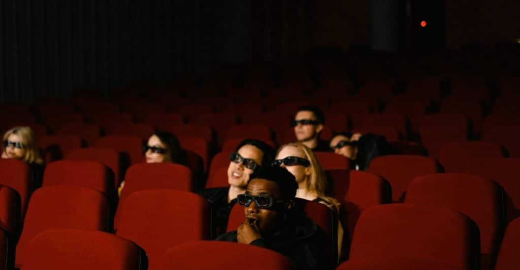 Several people sitting in red theater seats wearing 3D glasses, watching a movie in a dark virtual cinema.