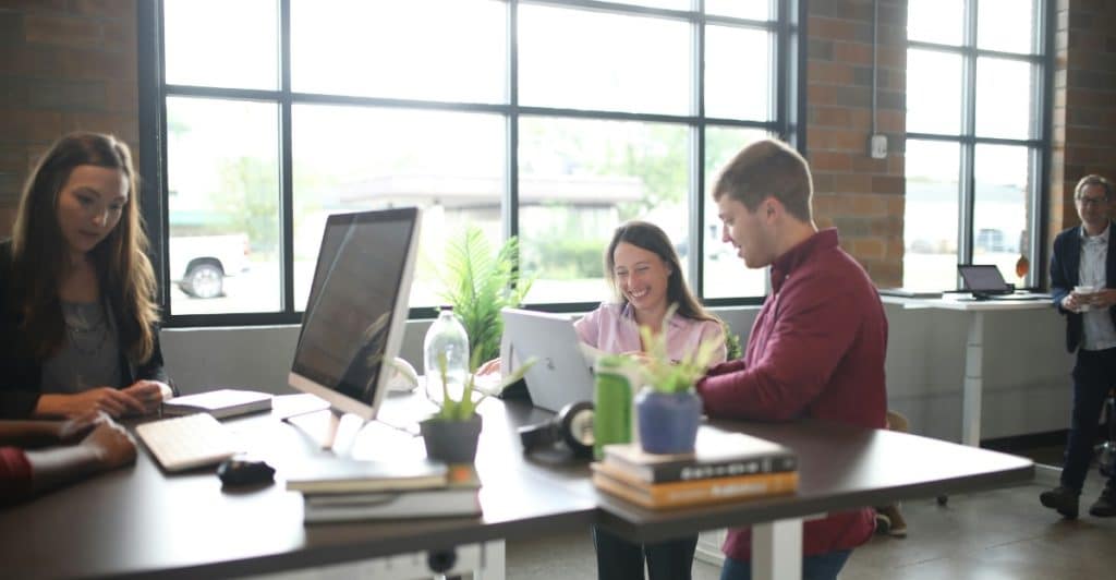 Several people working at standing desks in an open office environment with large windows