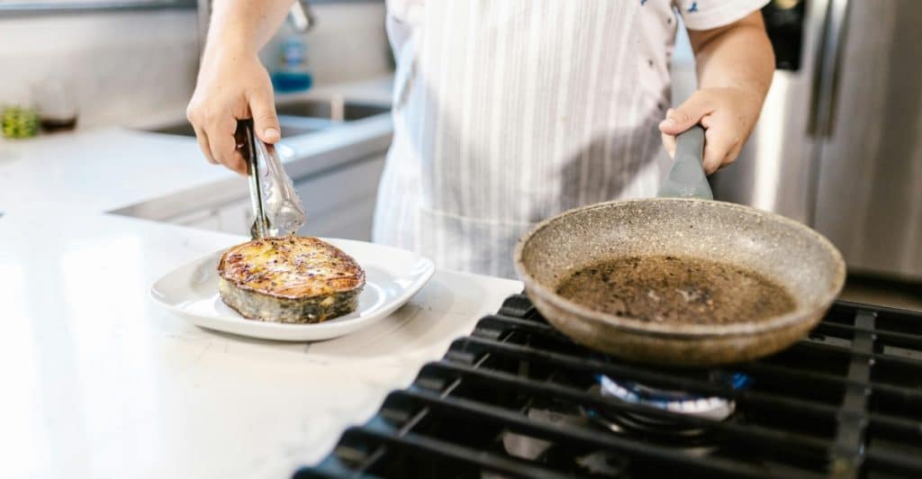 A person in a white apron using tongs to transfer a perfectly cooked piece of fish from a frying pan to a white plate in a kitchen.