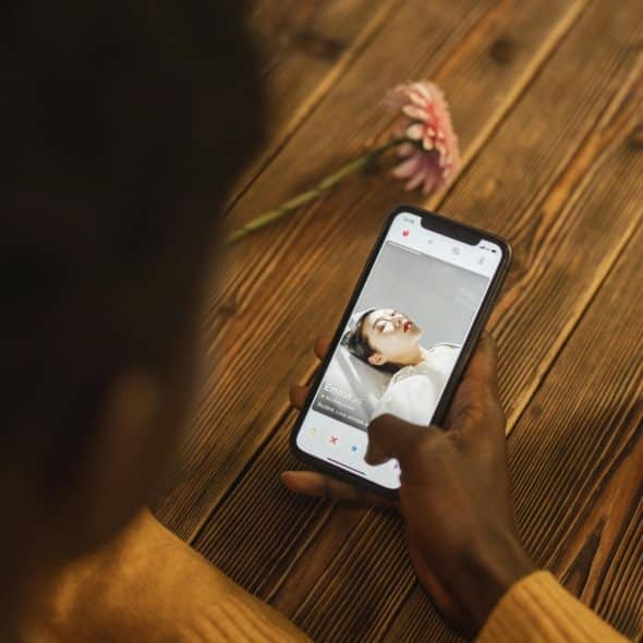 An overhead shot shows a person holding a black smartphone, actively scrolling through a dating app profile, with a white teacup and a pink flower visible on a dark wooden table beside them.