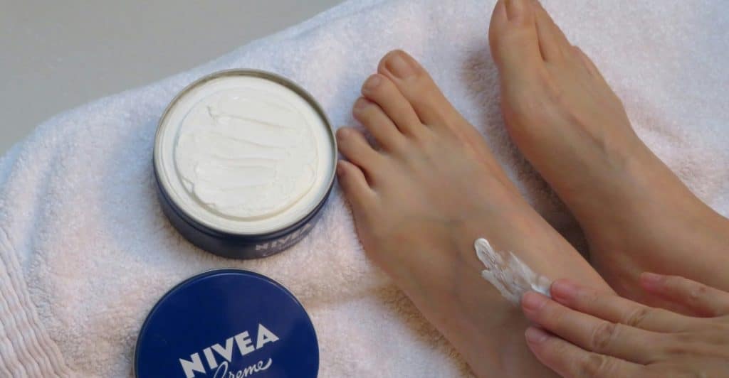 A person's hand applying white cream from a blue and white Nivea container to their foot, resting on a white towel.