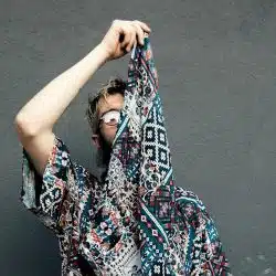 A person holding up a patterned shirt to obscure their face, standing against a dark wall.