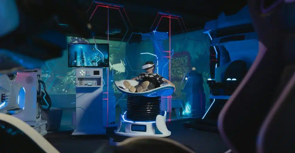 A person wearing a VR headset and sitting in a futuristic white lounge chair with blue glowing accents in a dark, digitally enhanced room.