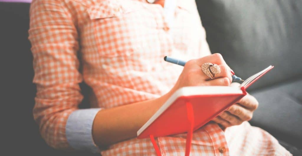 A person holding an open red journal and a pen, appearing to write.