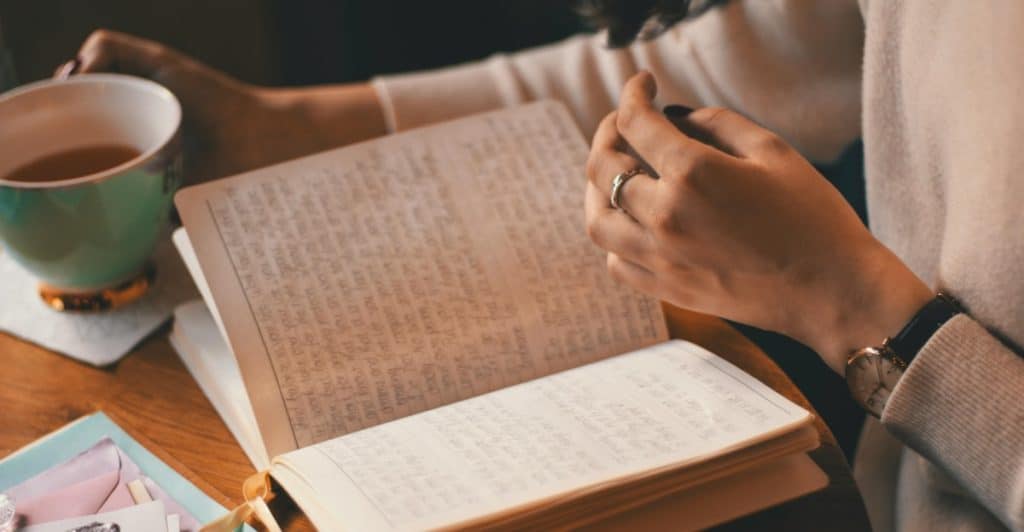 A person writing in a journal while holding a cup, with various stationery items on the table.