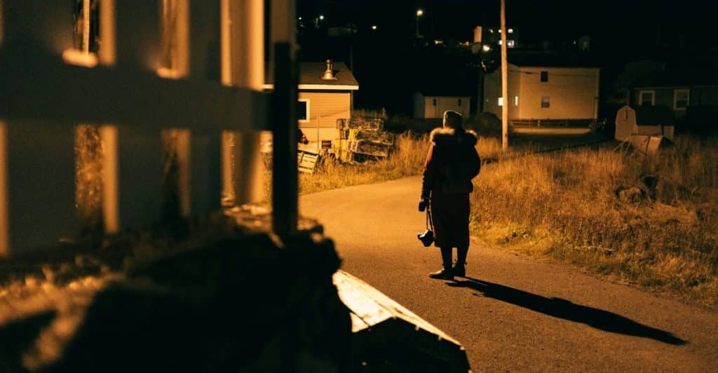 A person with a camera standing on a dark road at night, illuminated by a distant light.