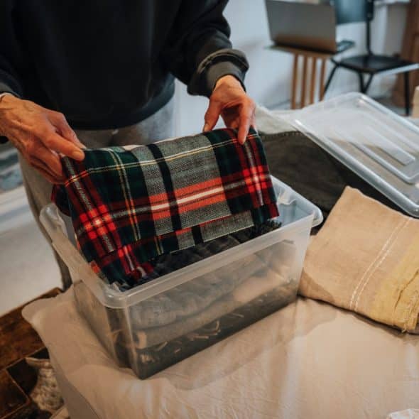 A person organizing folded clothes into a clear storage bin on a bed
